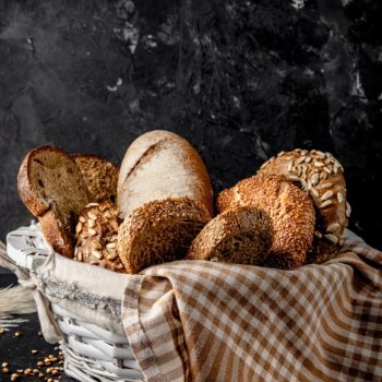 side view of basket full of breads as bagel baguette rye on black surface and black background